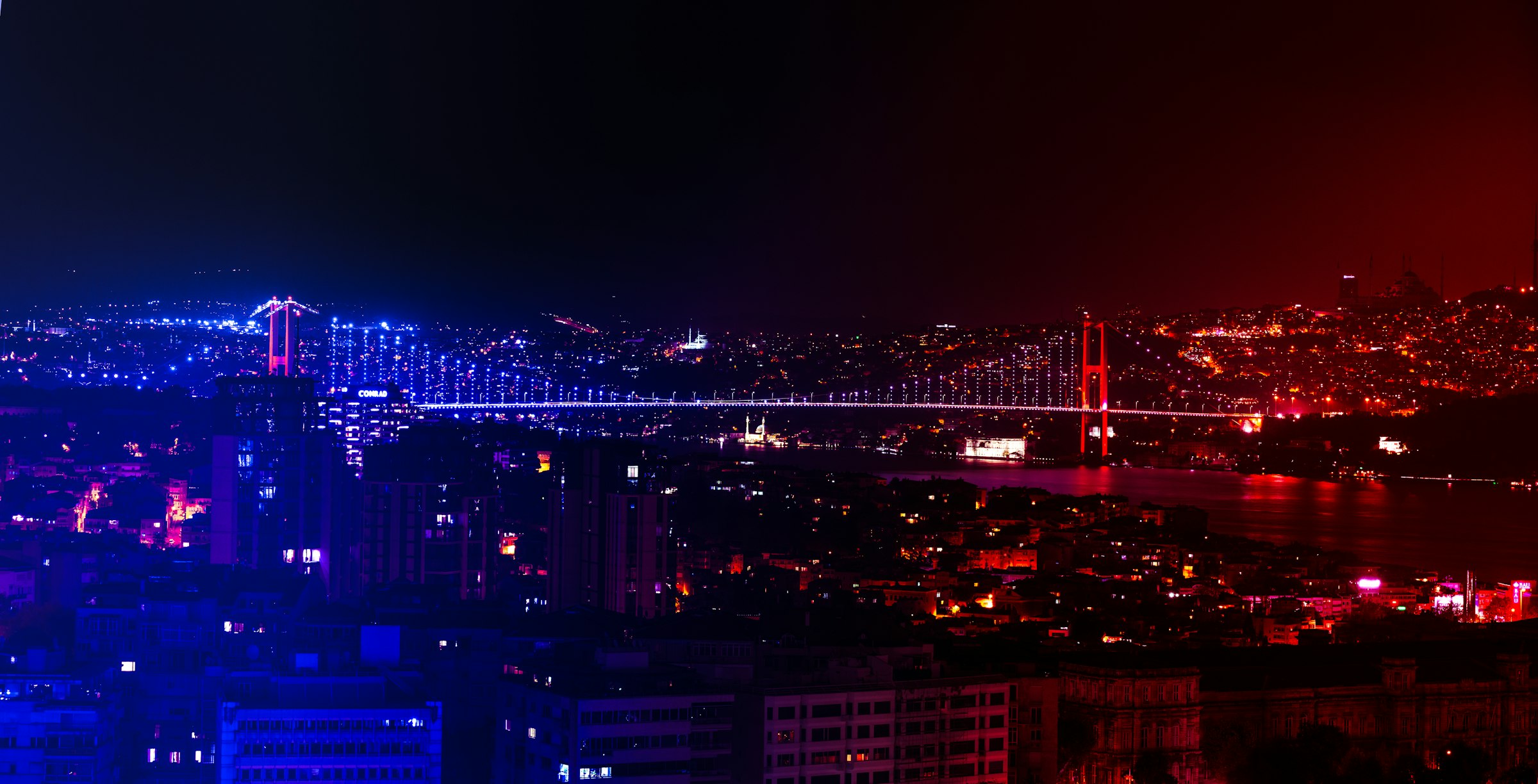 Aerial view of the Bosphorus Bridge lit blue and red over the strait at night